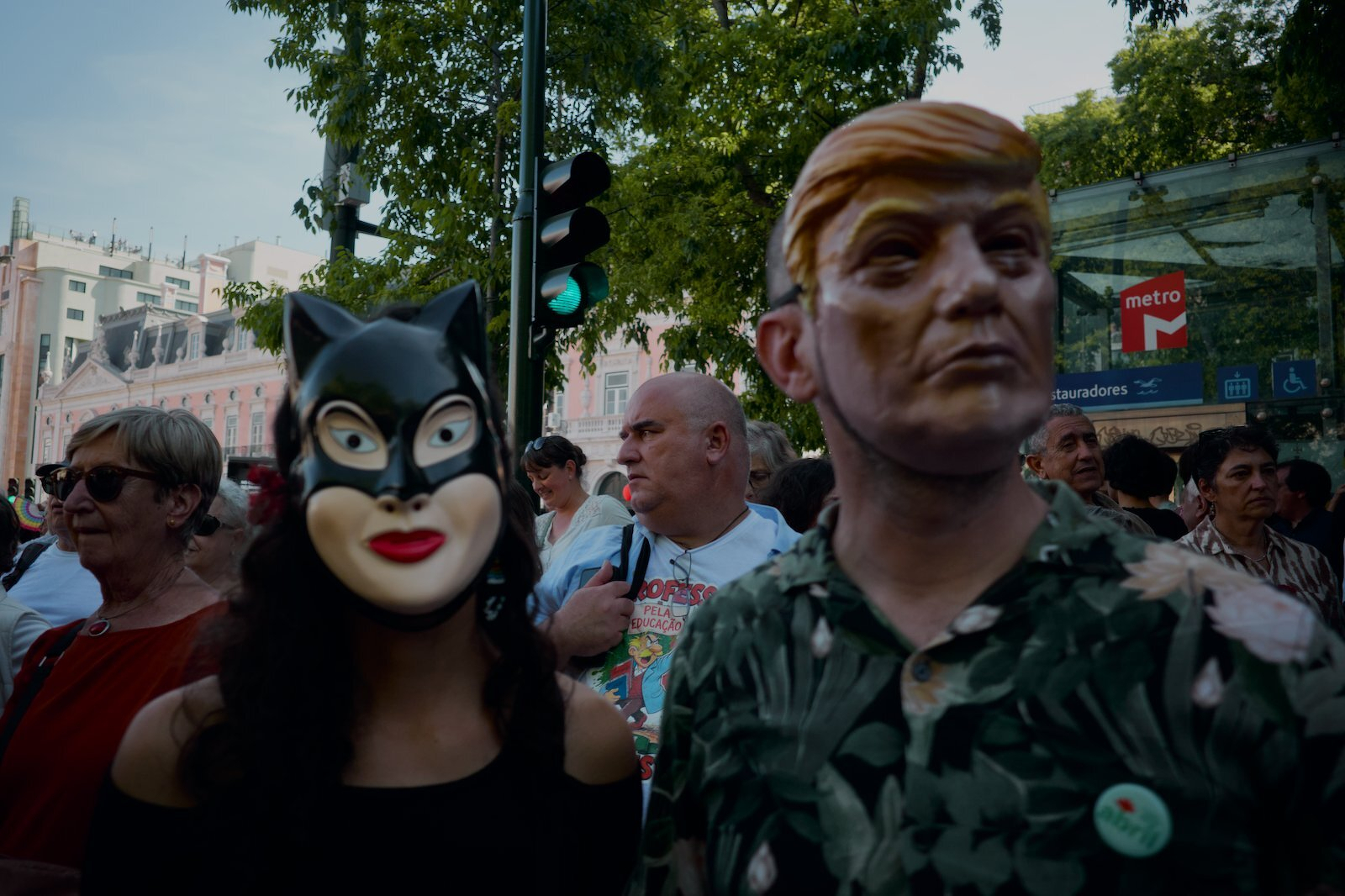 People wearing masks, including a cat-like mask and a mask resembling a famous figure, stand among a crowd near a metro entrance.