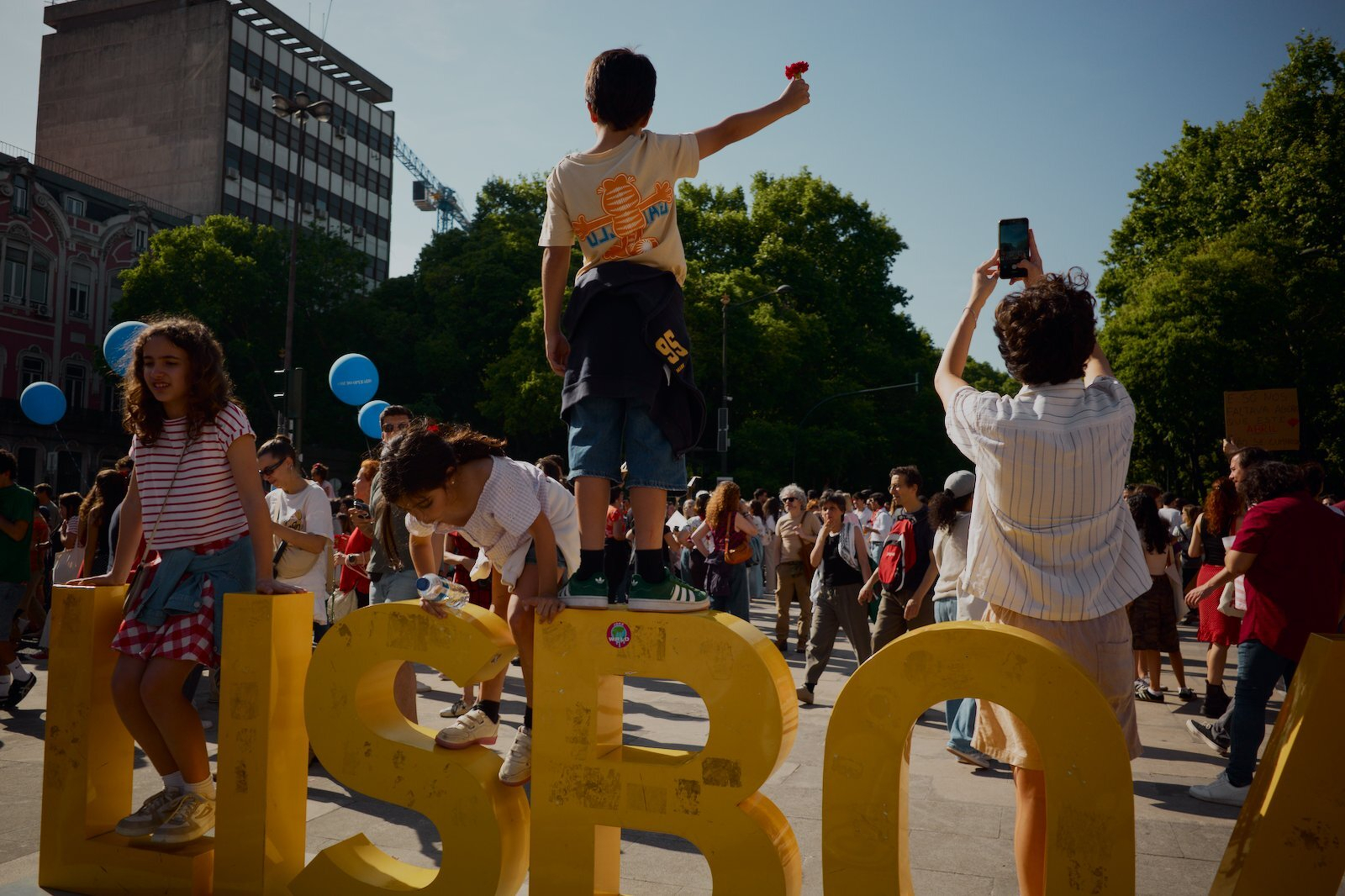People gathered at an outdoor event, with children climbing on large yellow letters spelling LISBOA and others taking photos.
