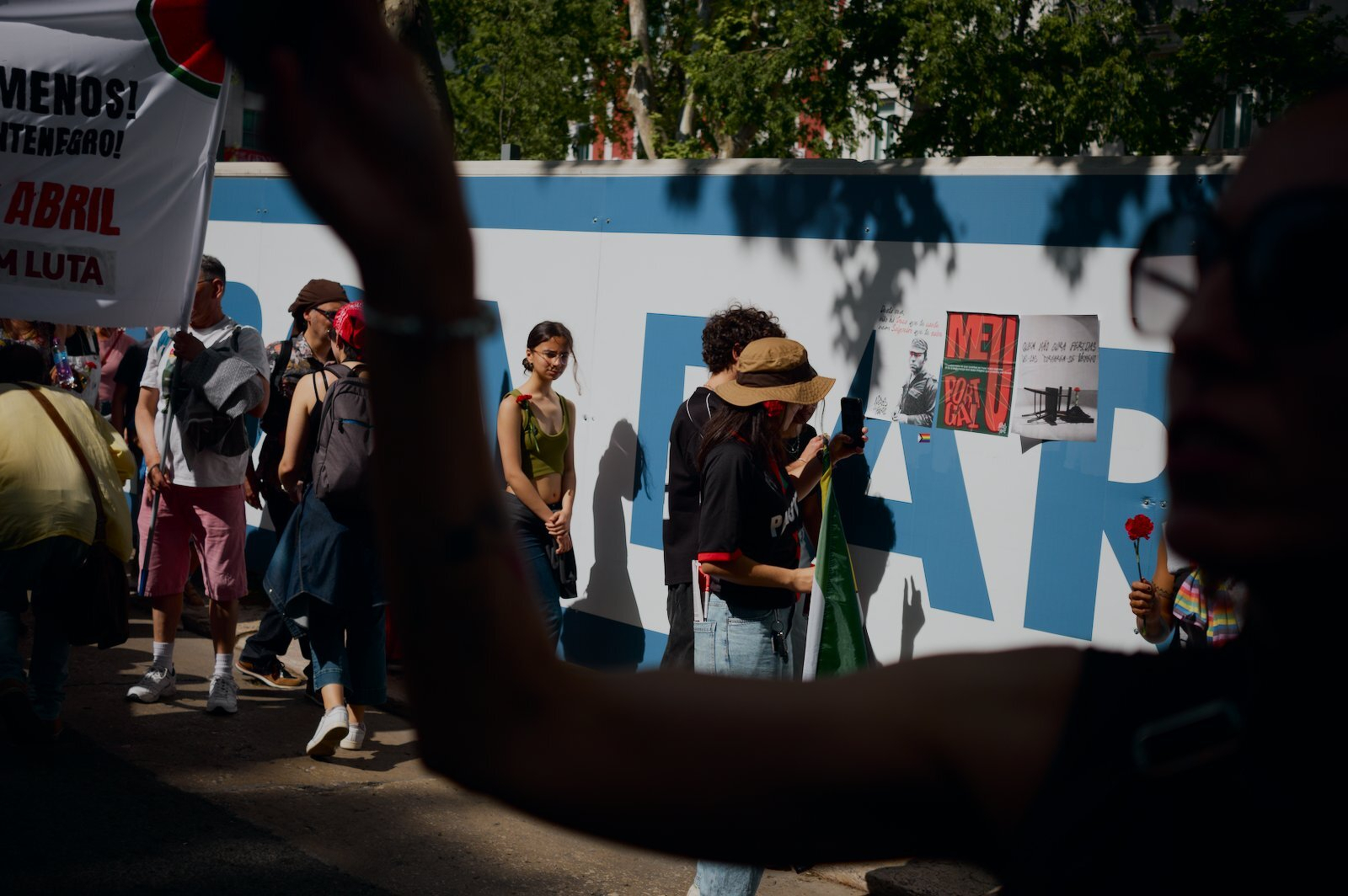 People are walking alongside a construction barrier, some holding signs and banners, with one person holding a red flower.