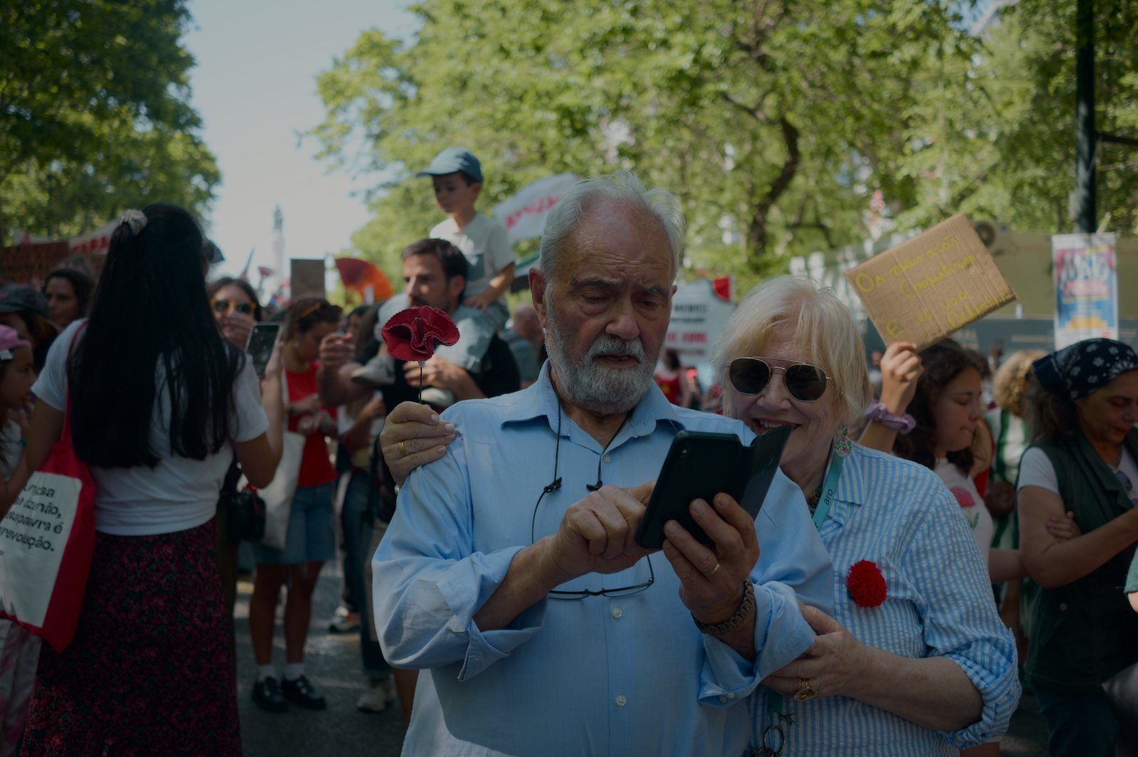 An older couple is looking at a smartphone surrounded by a group of people participating in a protest or demonstration, holding signs and wearing red.