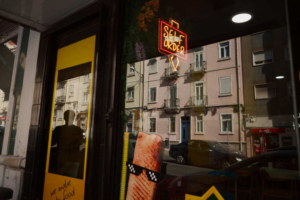 A storefront with a bright SELF ORDER neon sign reflects buildings and cars from across the street.