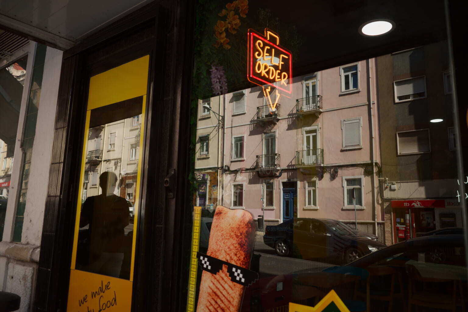 A storefront with a bright SELF ORDER neon sign reflects buildings and cars from across the street.