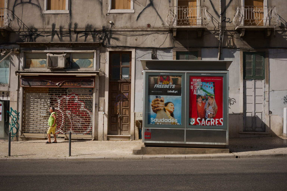 A man in a bright outfit walks past a graffiti-covered building and a bus stop with ads.