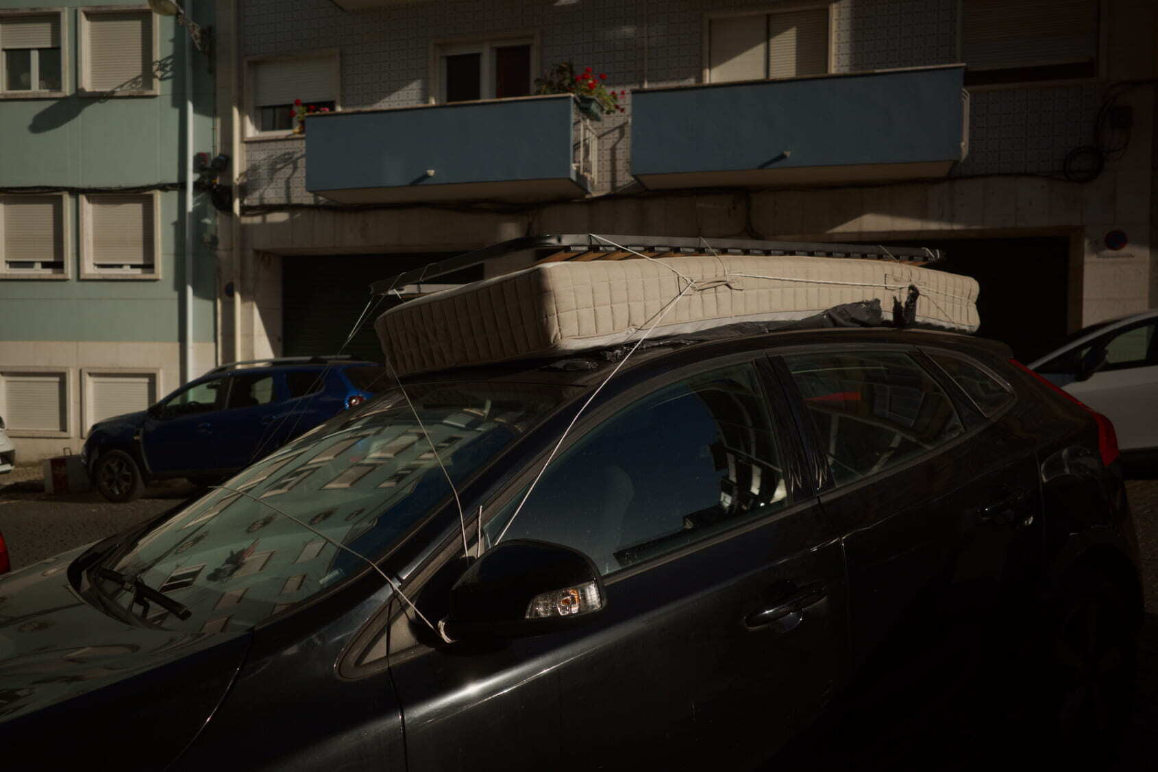 A mattress is secured on top of a car with cords in an urban setting.