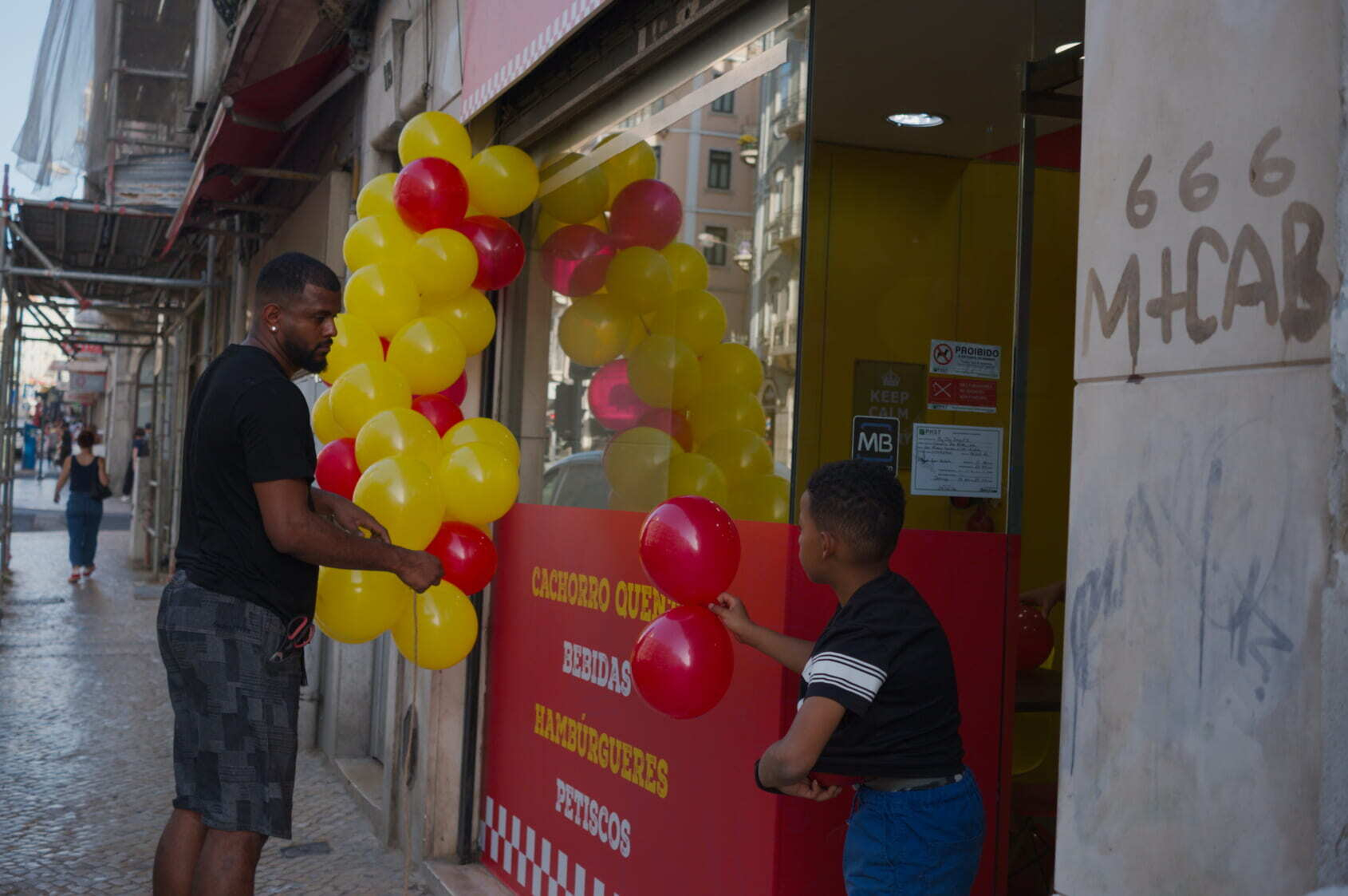 Two people are arranging yellow and red balloons at the entrance of a store with signs that include Cachorro Quente and Hamburgueres.