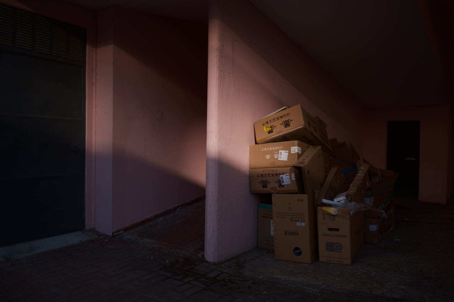 A stack of cardboard boxes and some scattered debris are placed against a pink wall in a dimly lit alleyway.