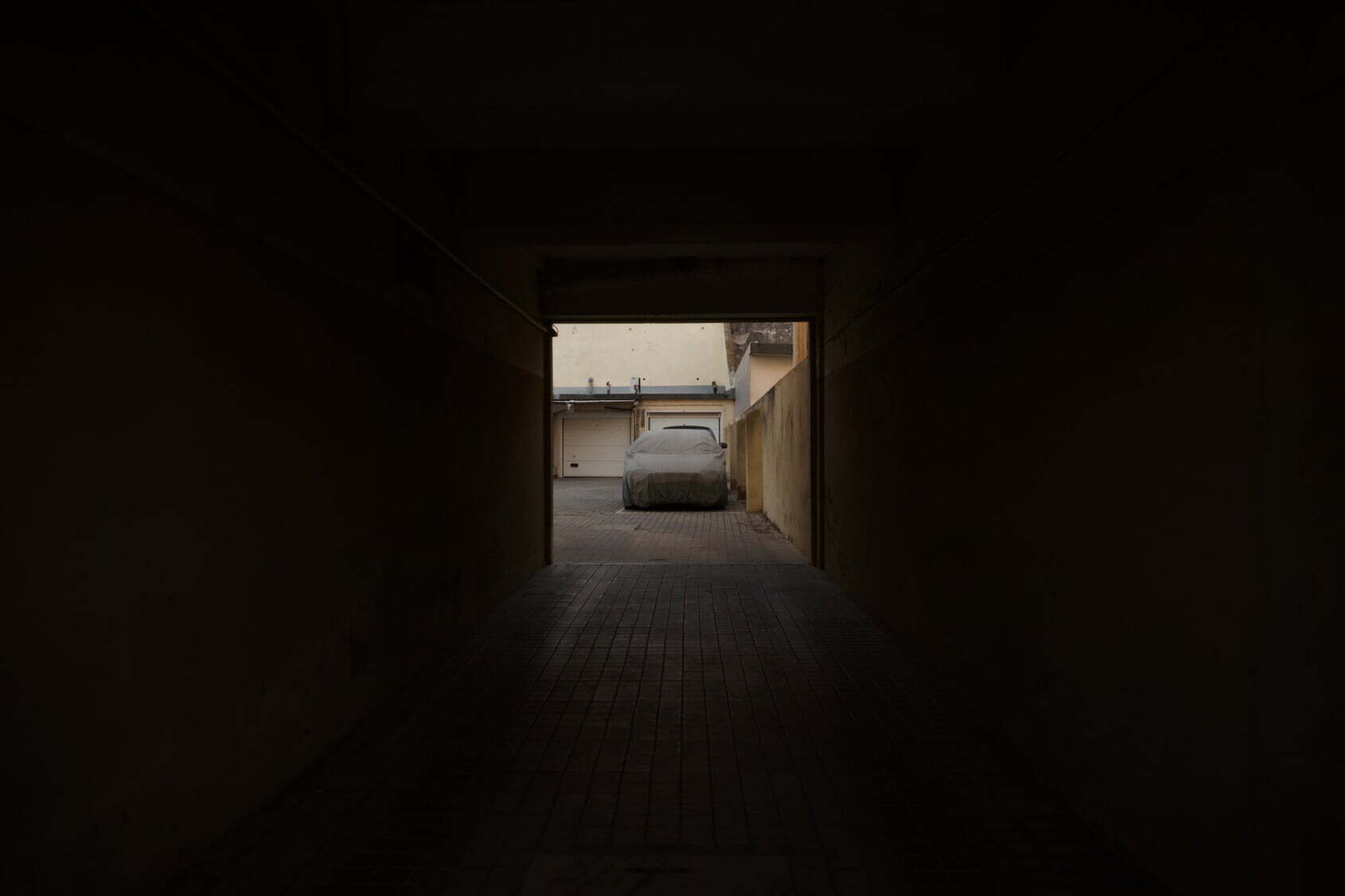 A dusty, old car is parked at the end of a dimly lit passageway leading to an outdoor area.