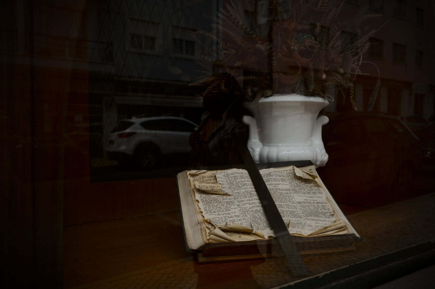 An old, worn book with yellowed and torn pages lies open beneath a white ceramic vase with a tall dried floral arrangement on a reflective glass surface.