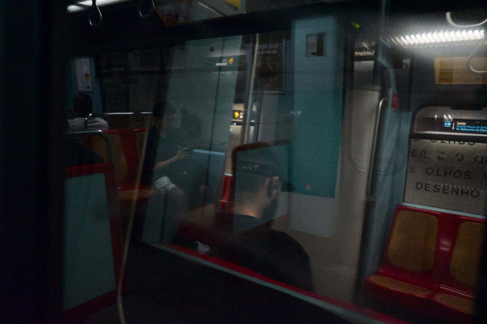 A person wearing a black cap is sitting inside a dimly lit subway train, with reflections on the glass adding a layered effect.