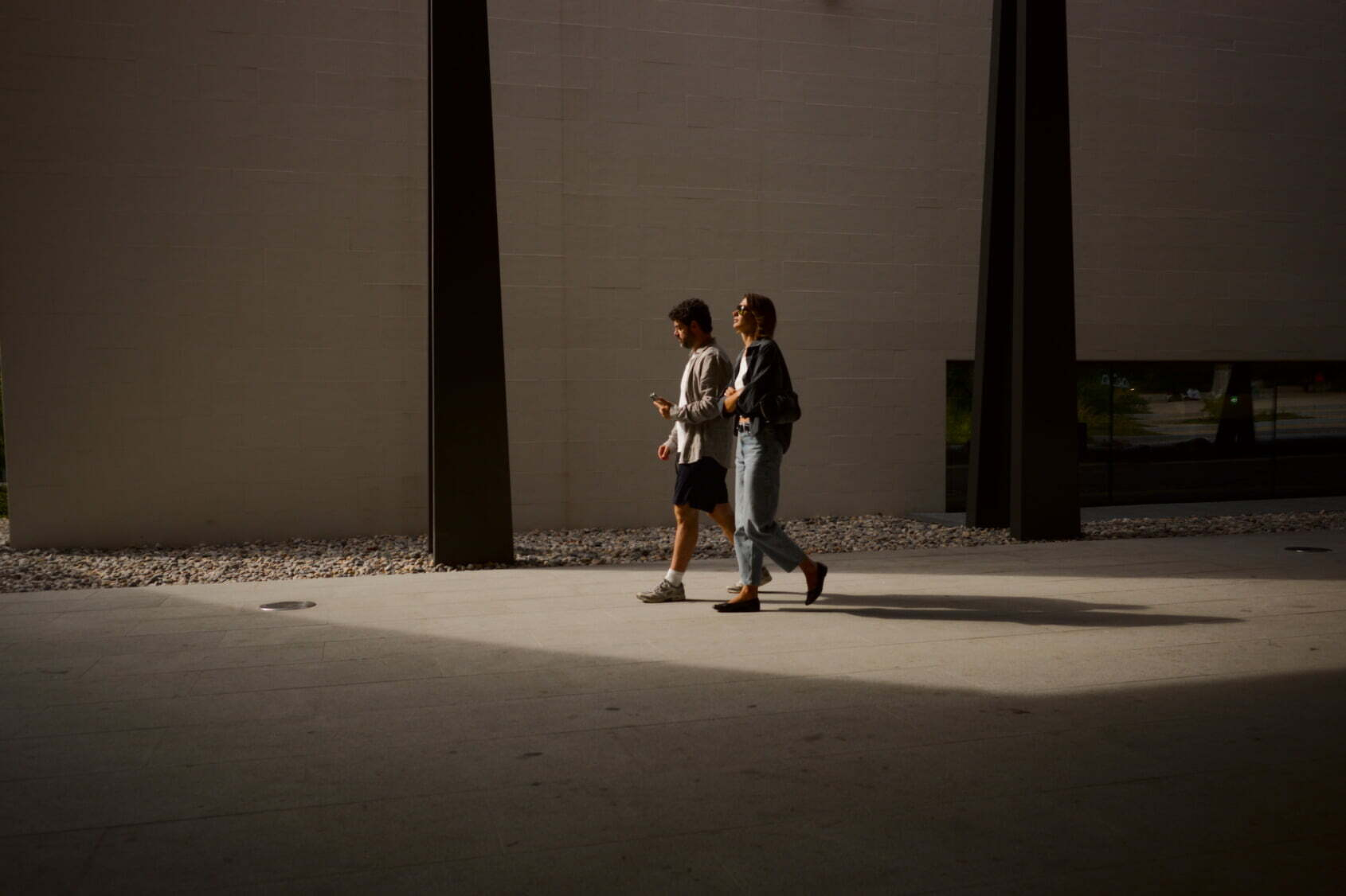 Two people are walking side by side through a sunlit area with long shadows cast on the ground.