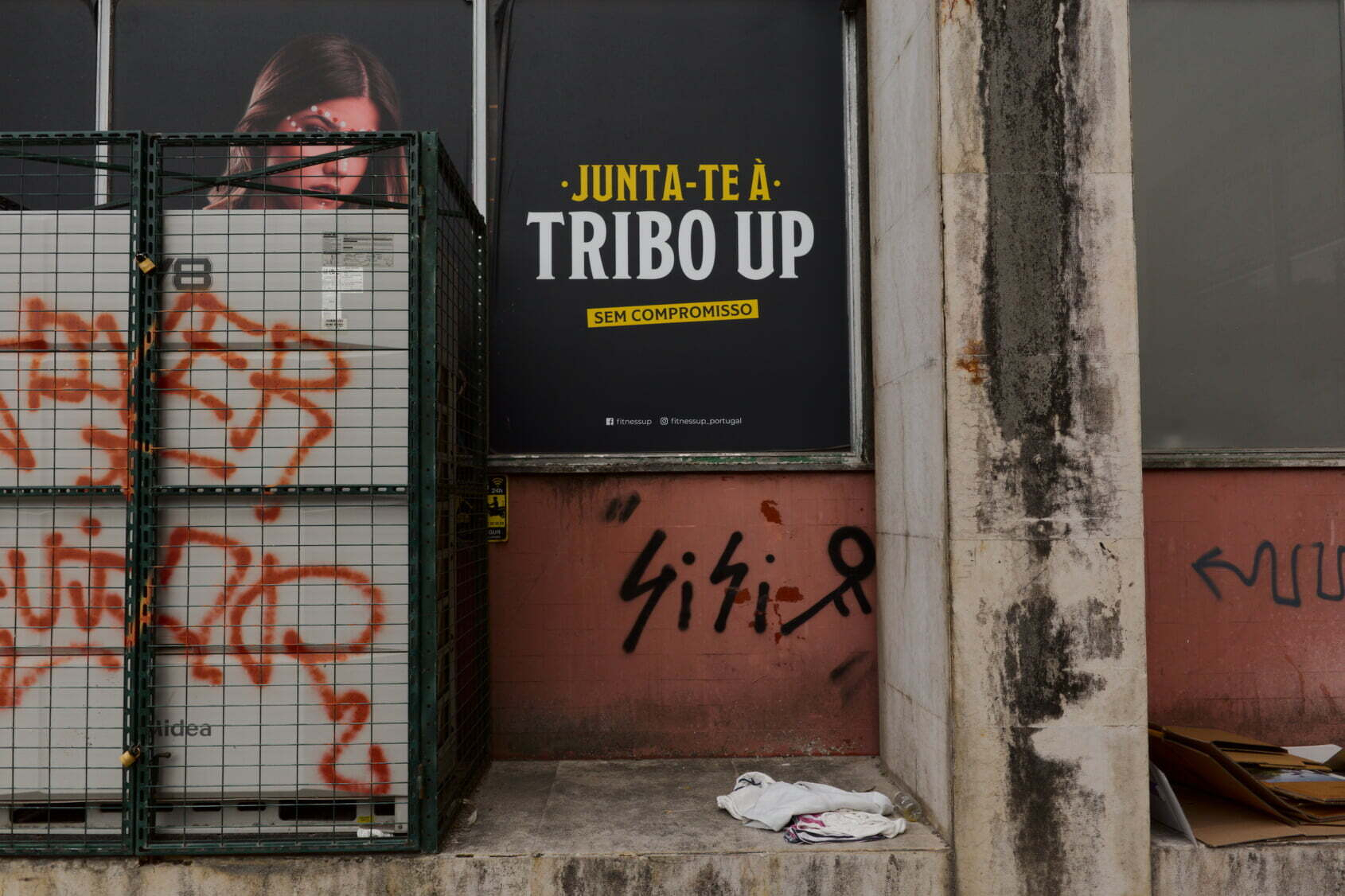 A wall displays a sign reading Junta-te à Tribo Up surrounded by graffiti and a metal fence with a partially obscured poster behind it.