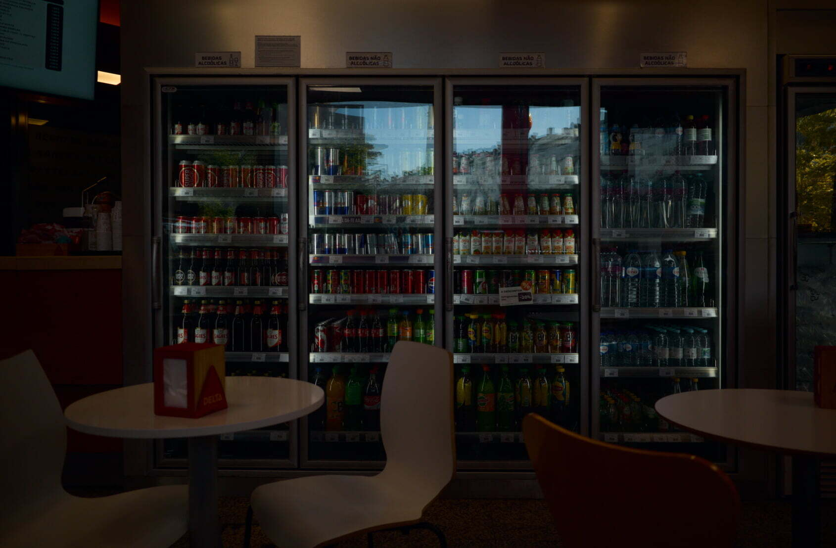 A refrigerated display case filled with various beverages is seen behind tables and chairs in a dimly lit cafe or restaurant.