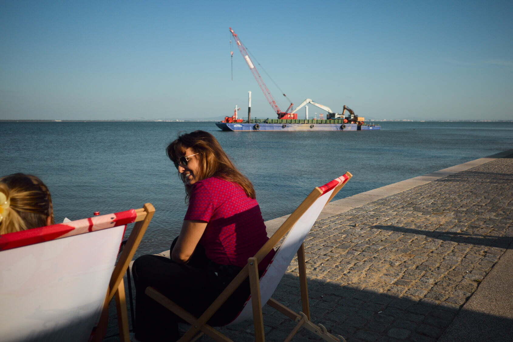 Two people are sitting on deck chairs near a waterfront while a crane on a barge operates in the water nearby.