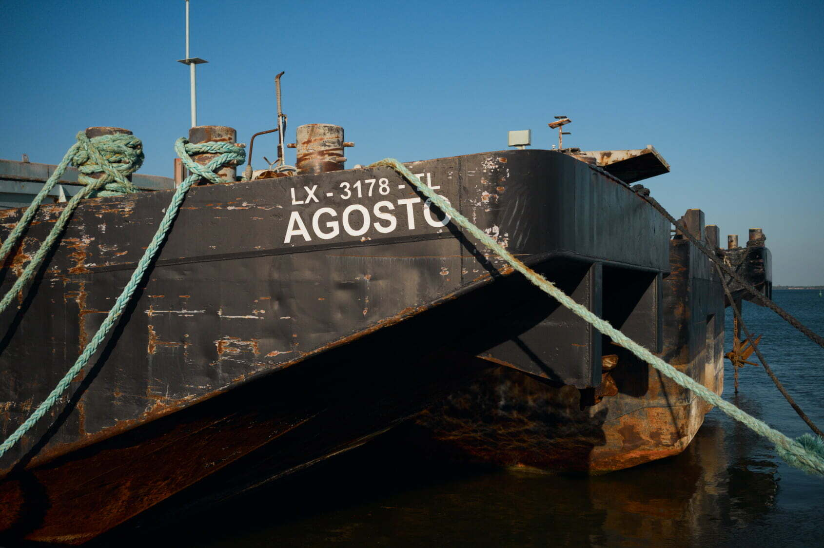 A rusted ship named AGOSTO with the identifier LX-3178-TL is tied to the dock with thick green ropes.