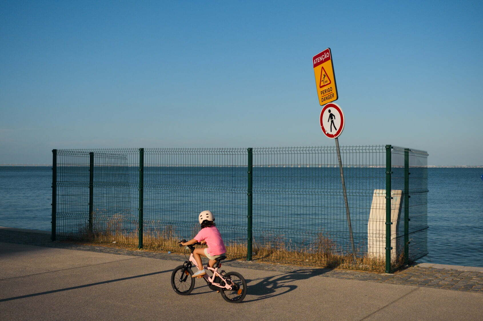 A child wearing a pink shirt and helmet rides a bike along a paved path next to a fenced area by the water with warning signs.