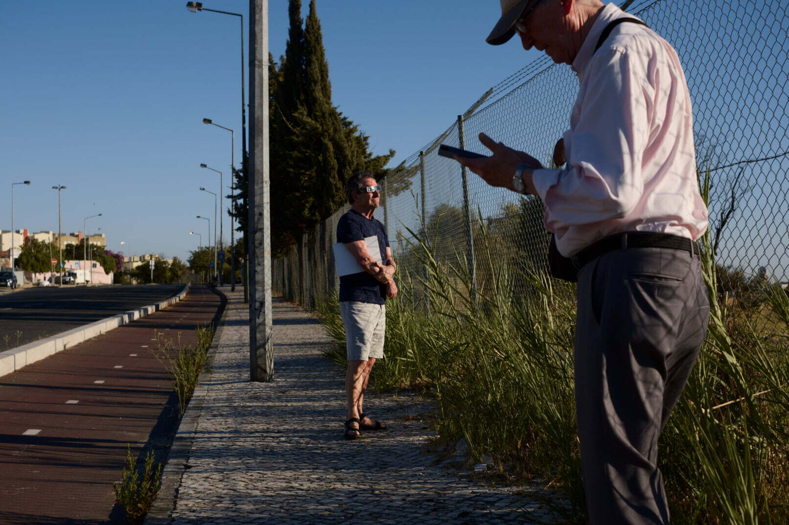 Two men are standing on a paved sidewalk next to a fence and vegetation, with one man writing or drawing in a notebook while the other looks on.