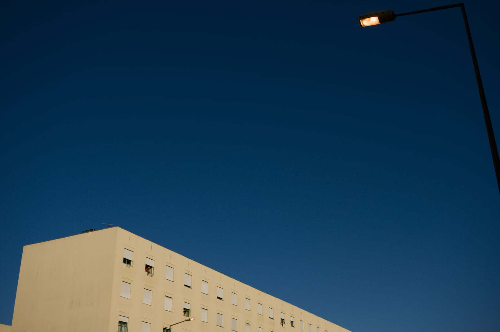 A beige apartment building with multiple windows is set against a clear dark blue sky, with a streetlight illuminated on the right side.