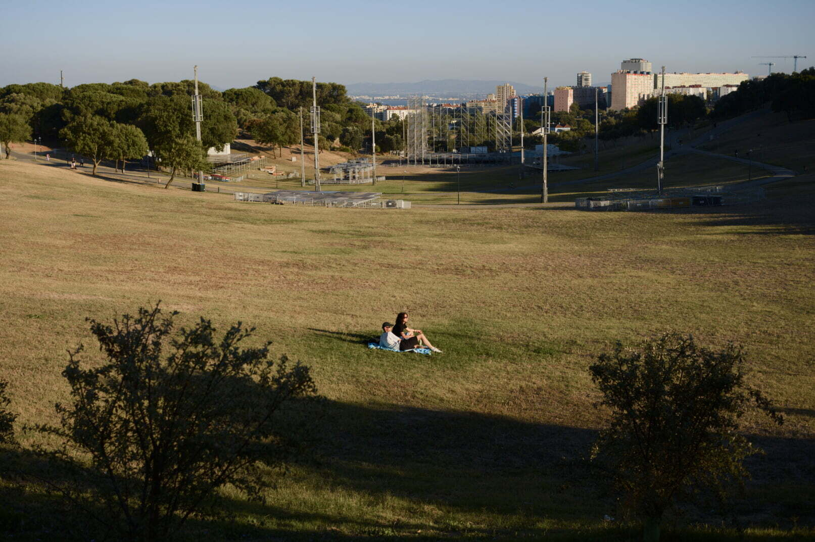 A couple is sitting on a grassy hill in a large open park with trees and buildings visible in the background under a clear sky.