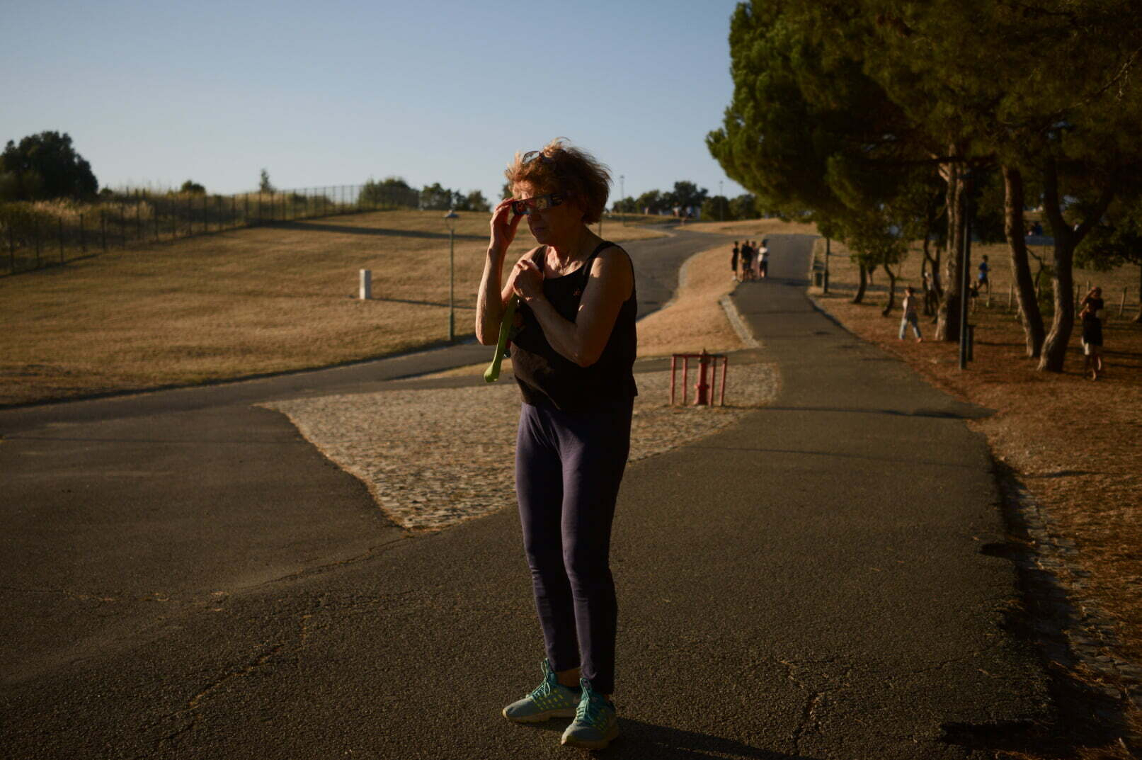 A person wearing athletic clothing and sneakers stands on a paved path in a park during a sunny day.