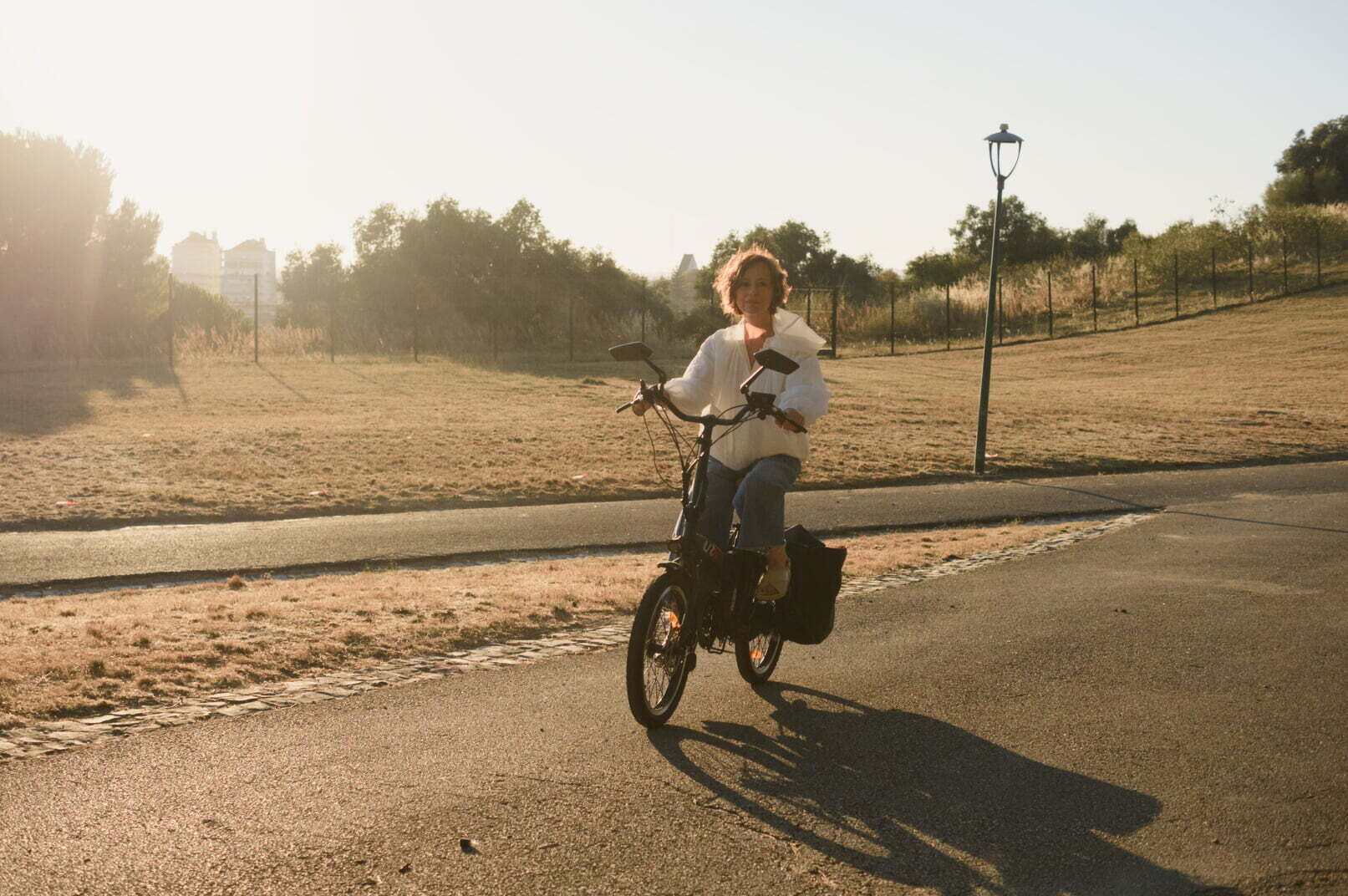 A person wearing a white shirt rides a bicycle on a paved path in a sunlit park during the late afternoon.