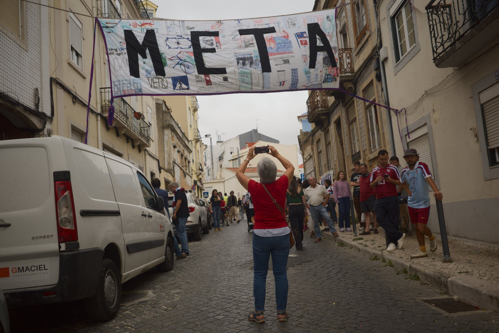 A person is taking a photo of a large banner with the word META hanging above a cobblestone street lined with buildings and observing people.