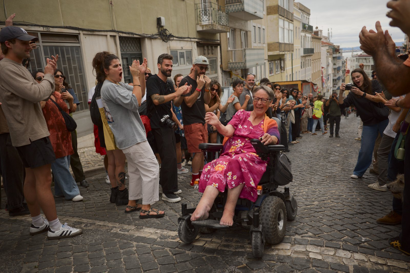 A woman in a wheelchair wearing a pink floral dress is smiling and moving along a cobblestone street while a crowd of people on both sides clap and cheer.