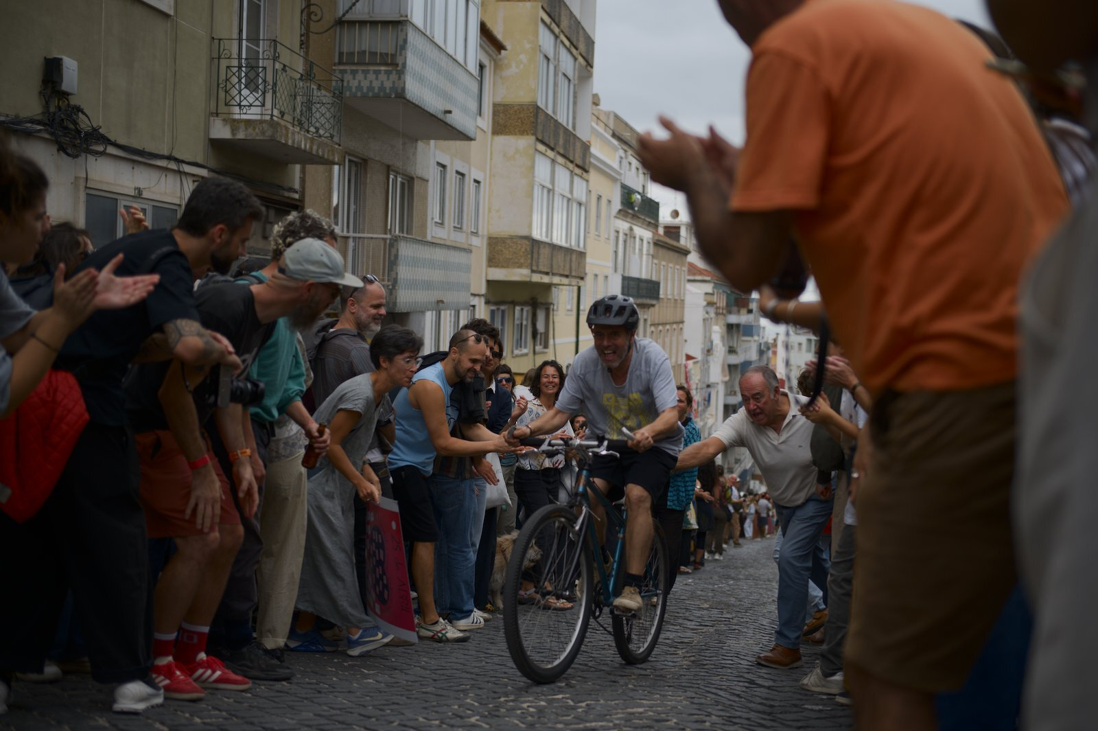 A cyclist wearing a helmet rides up a stone-paved street while a crowd of people on both sides cheer and applaud.