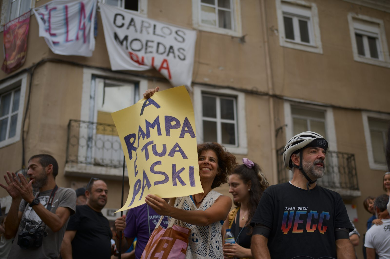 A group of people participates in a street protest, with one woman holding a yellow sign that reads A RAMPA É TUA PASKI while banners hang from the building behind them.