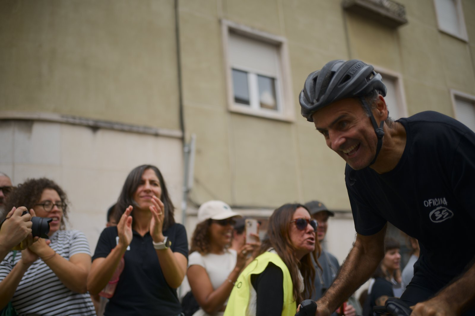 A man wearing a bicycle helmet is smiling while a group of people in the background clap and take photos.