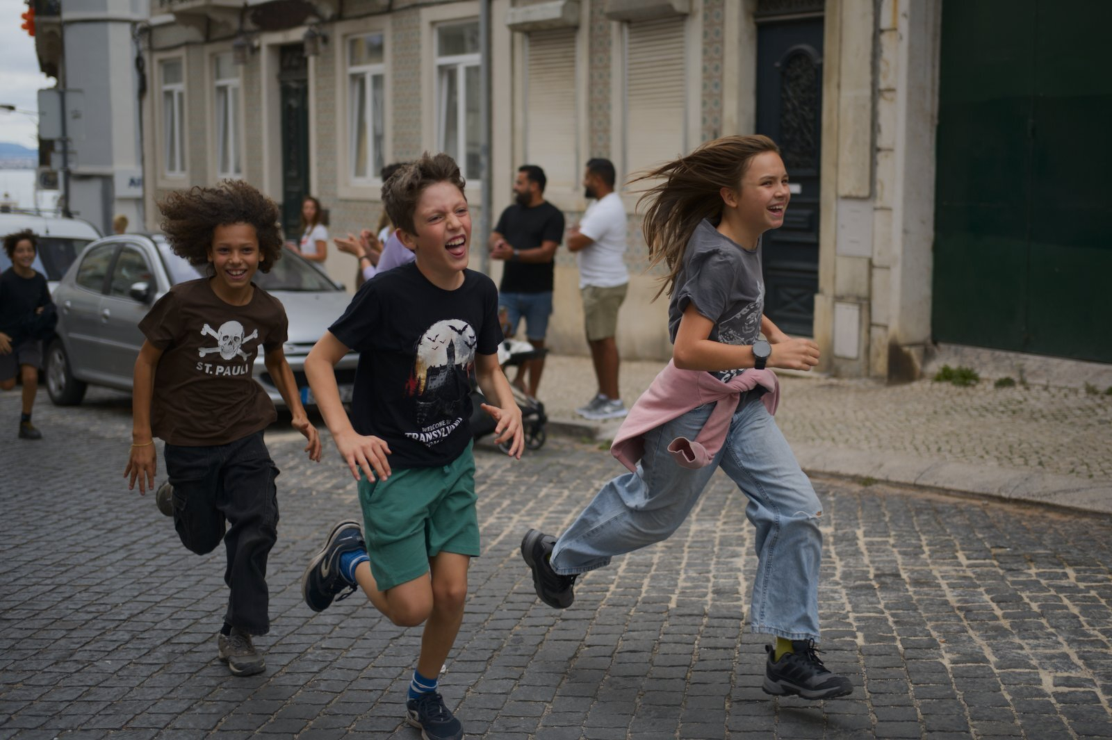 Three children are joyfully running down a cobblestone street in a lively urban area.