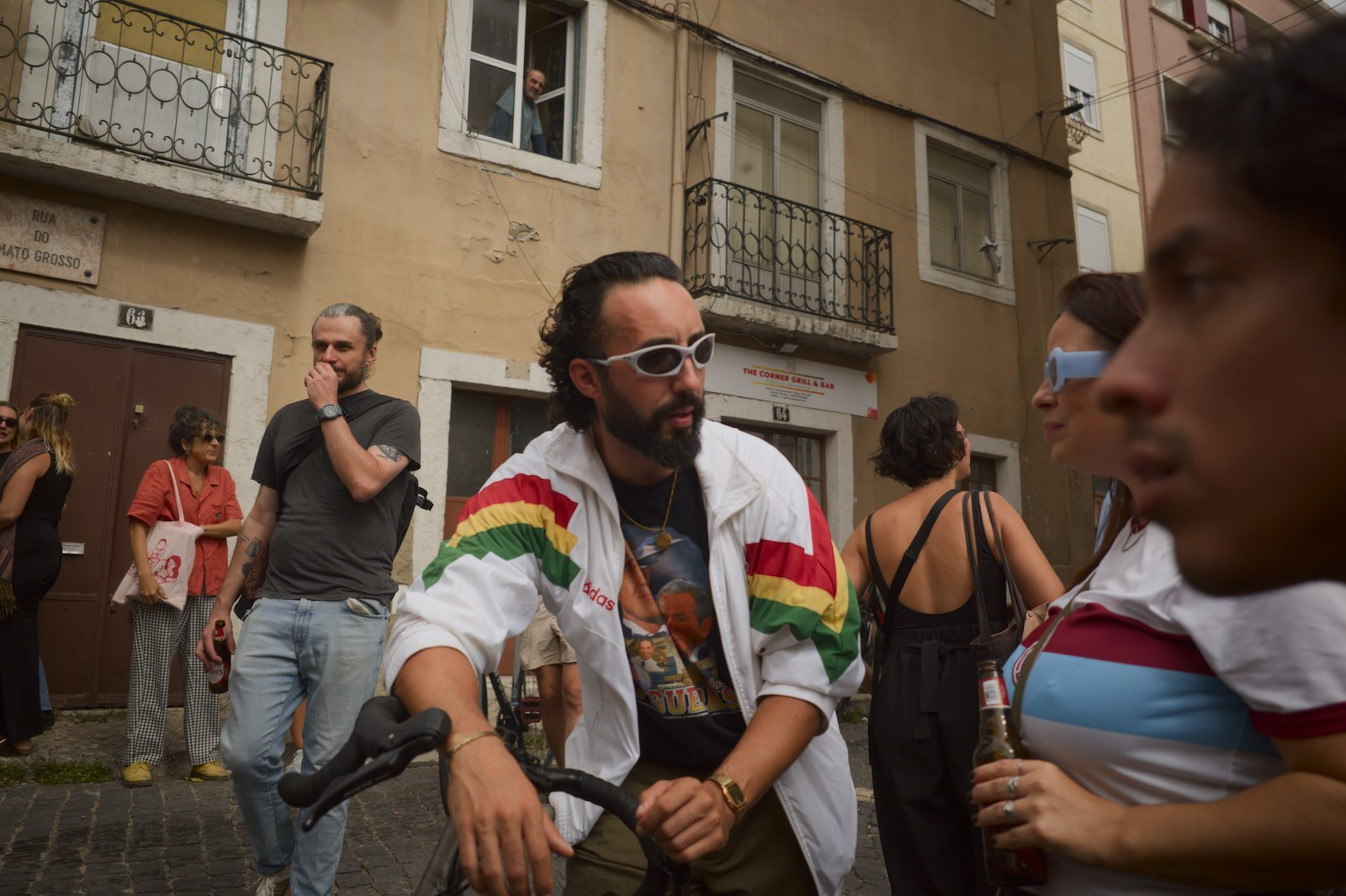 A group of people are gathered outdoors on a street, with one man in a white jacket with colorful stripes holding a bicycle and wearing sunglasses.