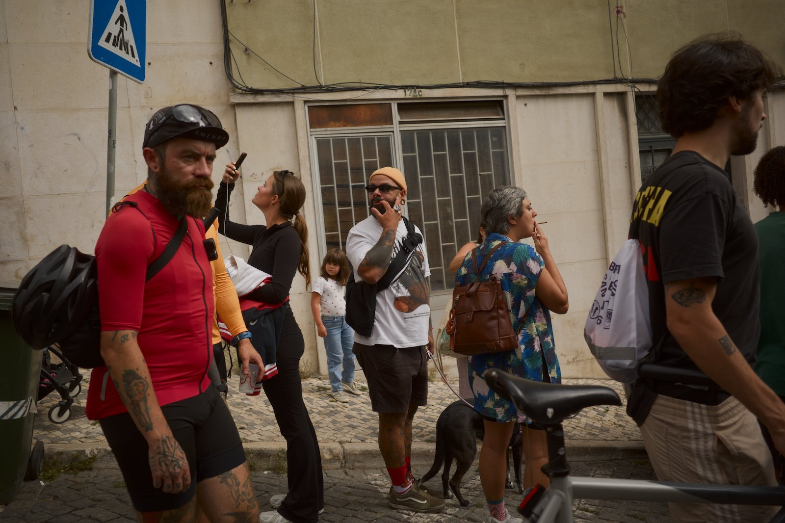 A group of people, including a cyclist in red gear, are gathered on a street, some looking at their phones while others are engaged in conversation or standing nearby.