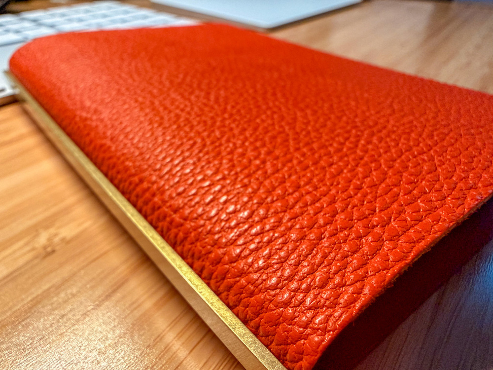 A red leather-bound item, possibly a book or journal, with a textured cover and a gold-colored edge, rests on a wooden surface near a keyboard.