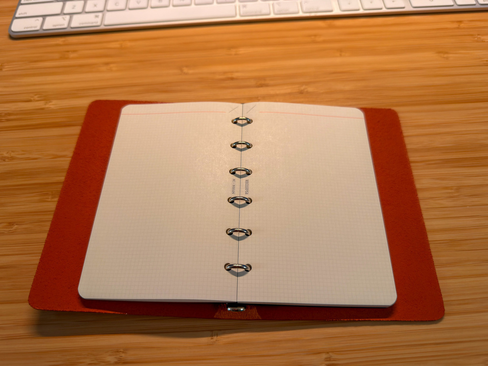 An open notebook with grid paper and binder rings rests on a wooden desk beneath a keyboard.