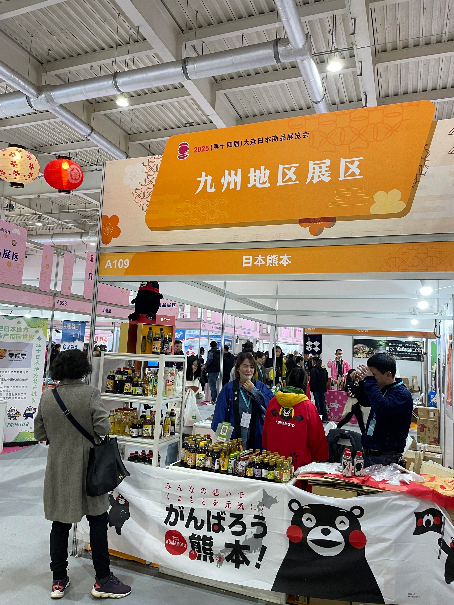 People browsing and interacting at a booth showcasing products from the Kyushu region, featuring merchandise and decorations with a bear mascot.