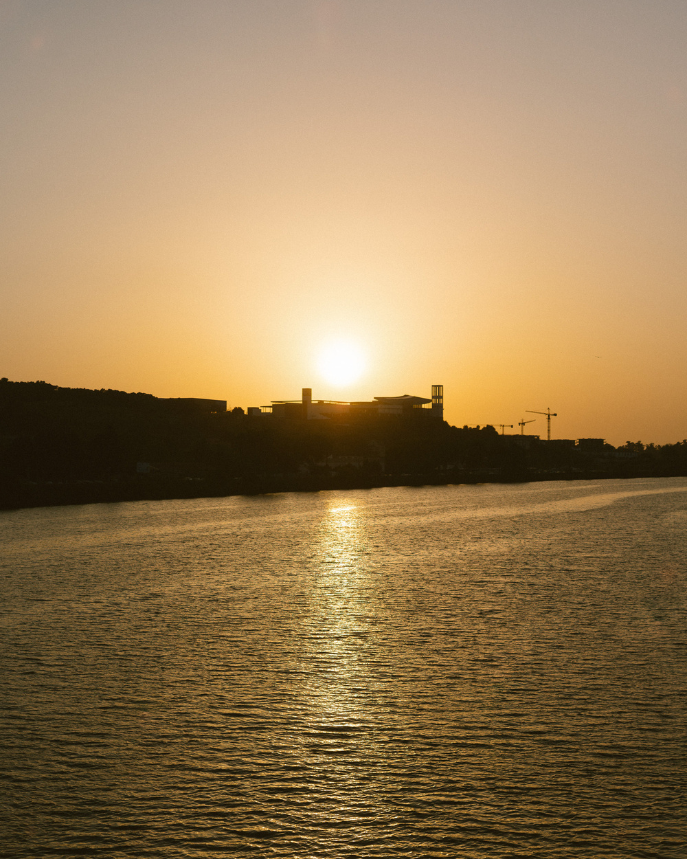 A serene sunset casts a warm glow over a body of water with silhouettes of buildings and cranes in the distance.