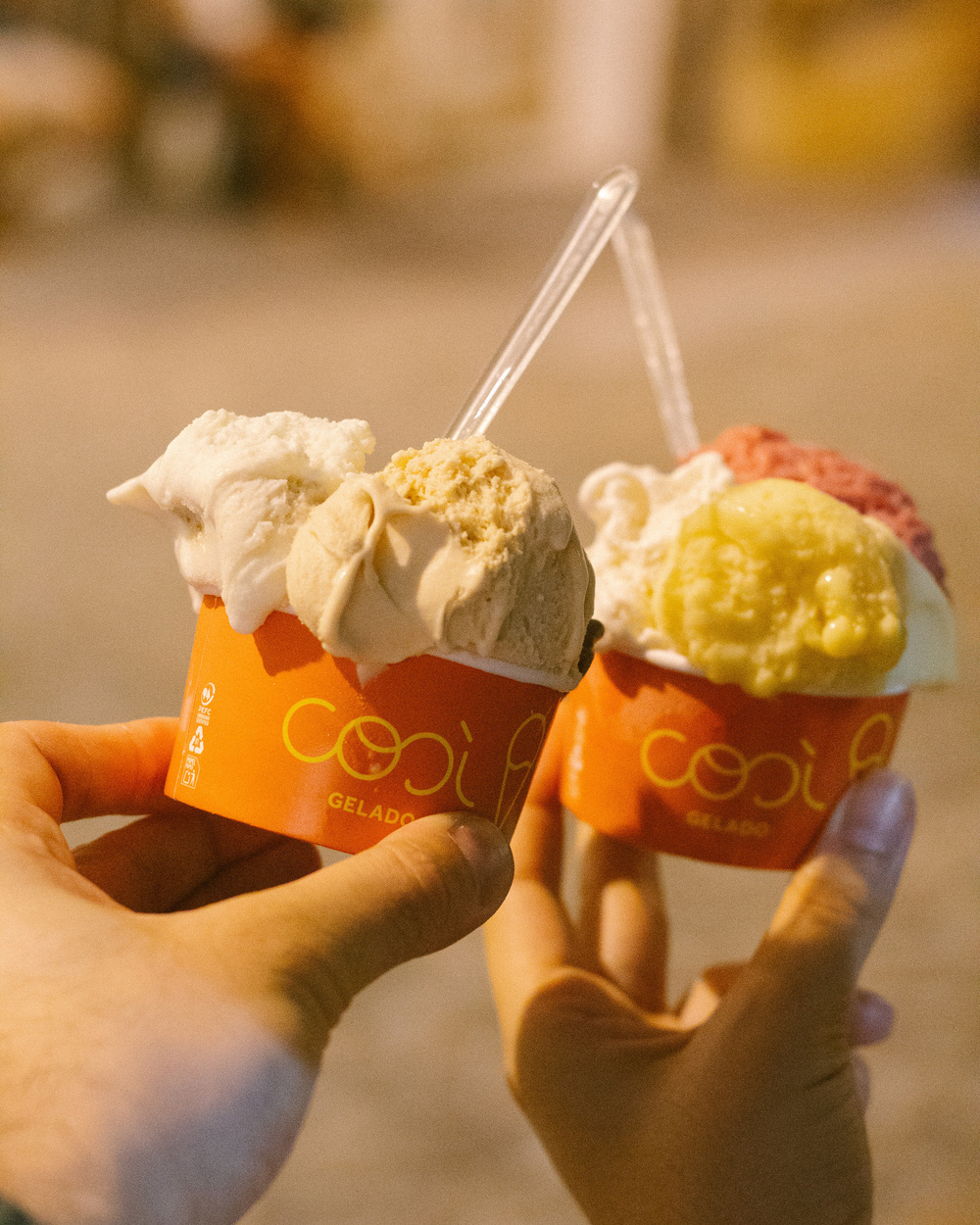 Two hands hold cups of ice cream with multiple flavors, each topped with a small spoon, in a warmly lit setting.