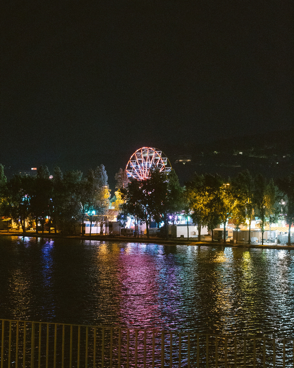 A ferris wheel and colorful lights illuminate the night sky by a riverside, reflecting on the water.