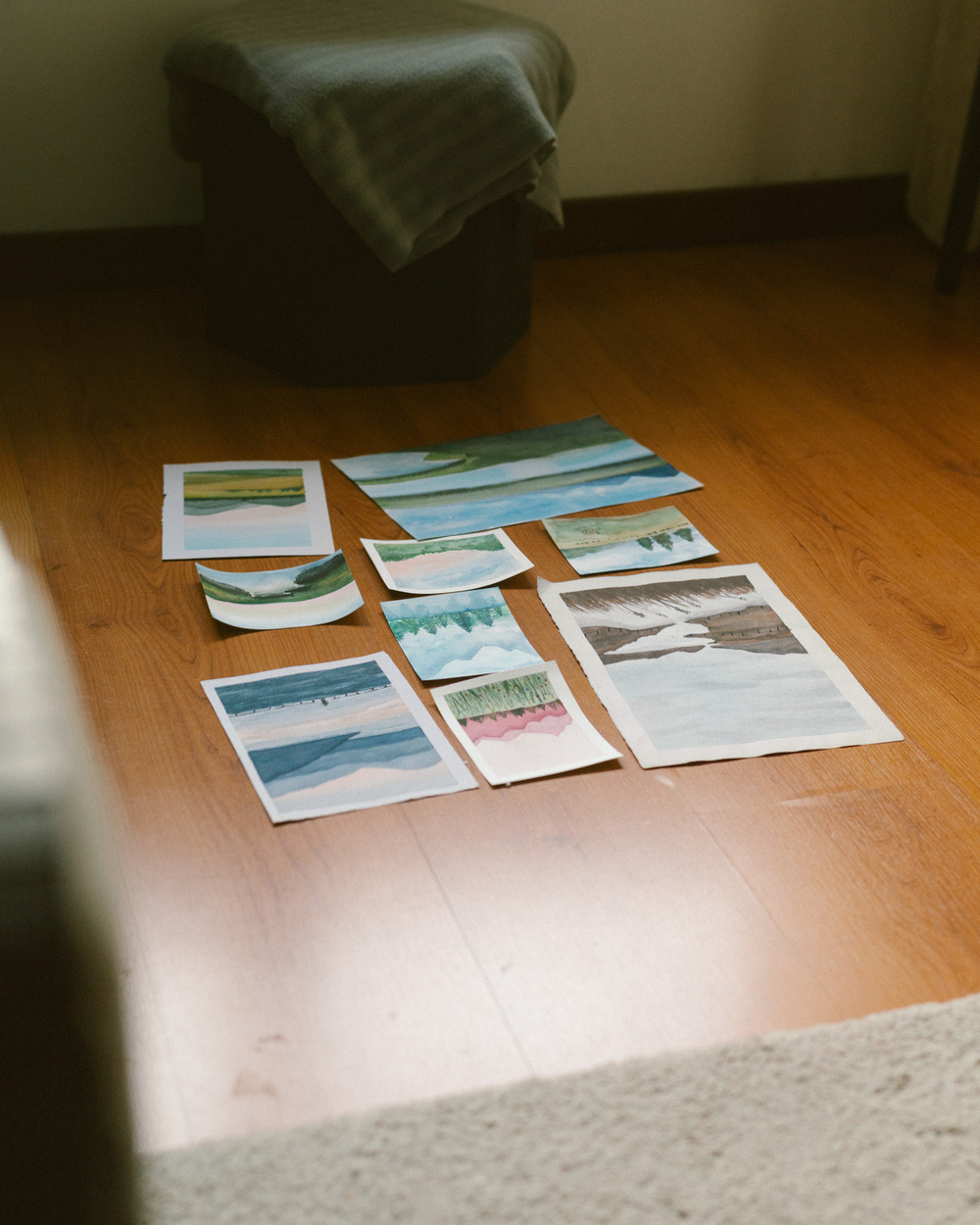 A collection of landscape paintings is arranged neatly on a wooden floor next to a covered piece of furniture.
