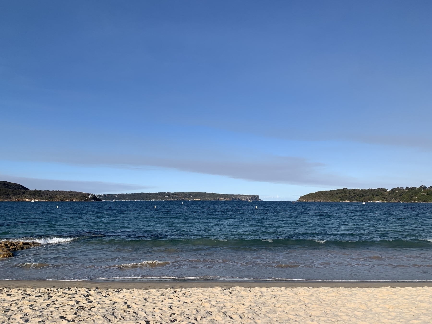 Photo of Middle Harbour, Sydney taken from Edward’s Beach facing towards the gap between Middle Head (on the right) and North Head. Blue skies over the harbour with thin strips of dirty-grey clouds.
