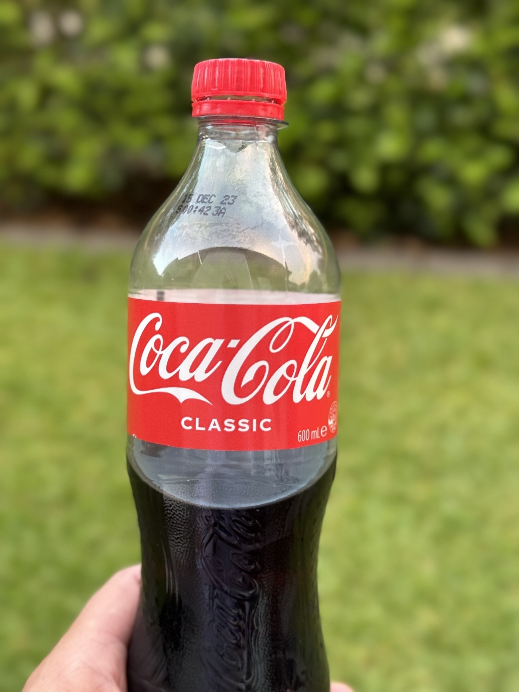 A half-finished bottle of Coca Cola in front of a lawn and a hedge