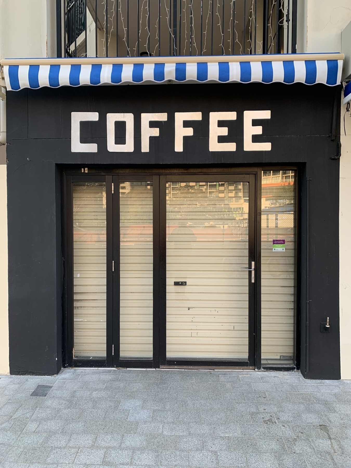 Photo of a closed shop front with glass walls (in front of a closed roller shutter) with a large “COFFEE” sign above the entrance