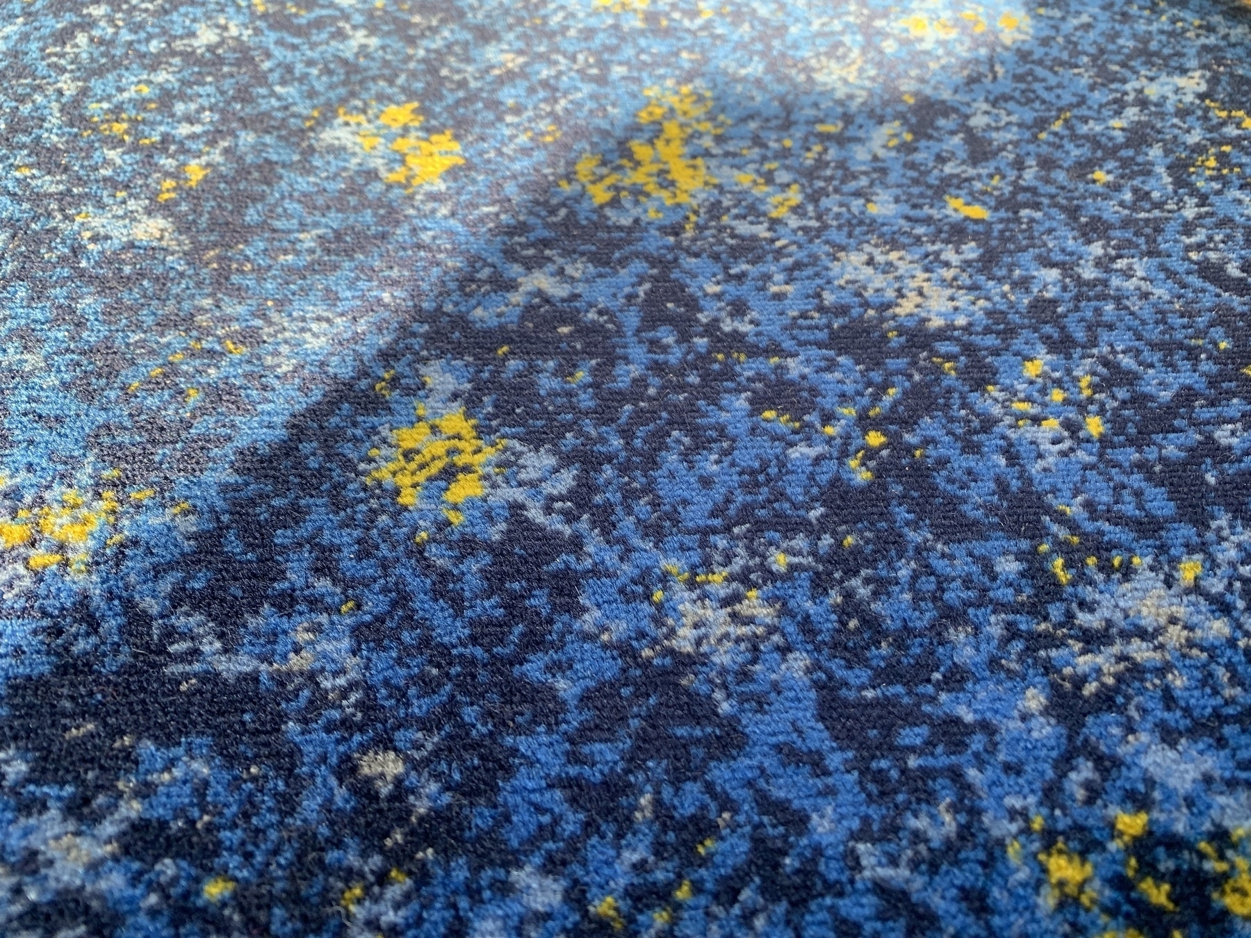 Fabric train seat cover in a mottled design mixing 4 shades of blue and some yellow, partially in shadow, partially in bright sun.