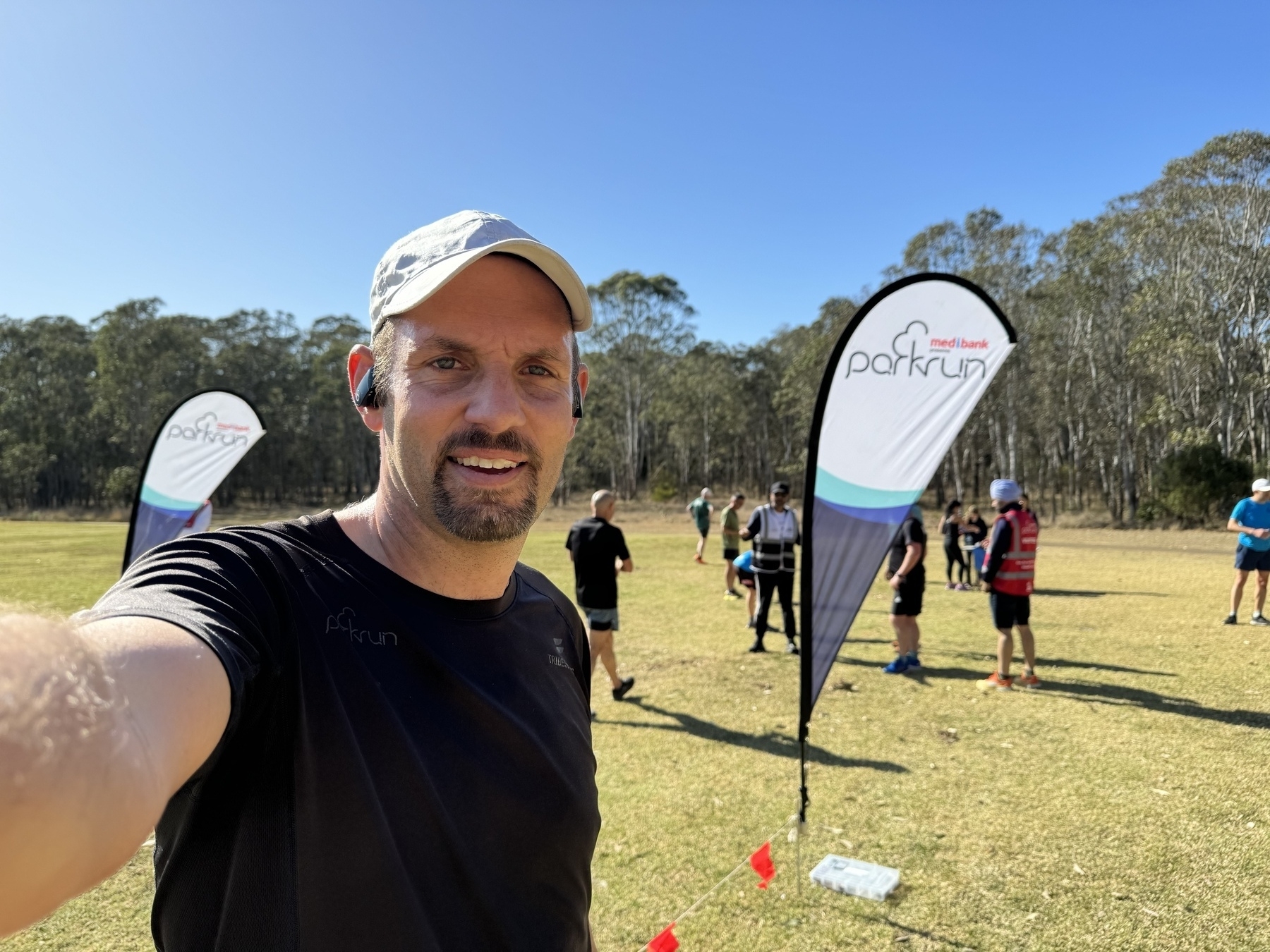 A selfie of me (41 year old male with a goatee wearing black running clothes and a white running cap) standing in front of Parkrun banners and other parkrun participants and volunteers