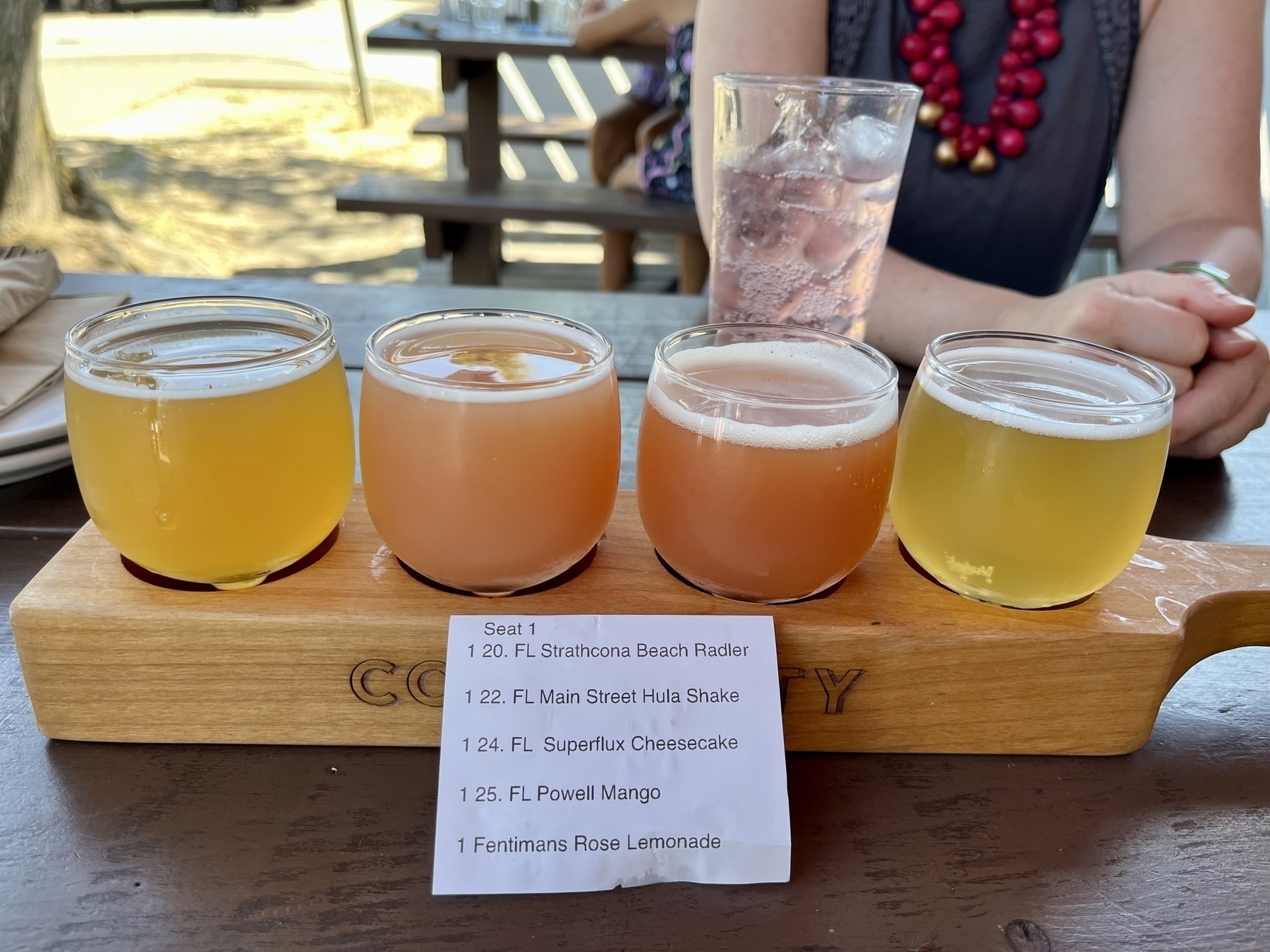 Flight of 4 beer tasters in shades of orange and pink - with a print out listing them &10;&10;Strathcona Beach Radler&10;1 22. FL Main Street Hula Shake&10;1 24. FL Superflux Cheesecake&10;1 25. FL Powell Mango&10;1 Fentimans Rose Lemonade