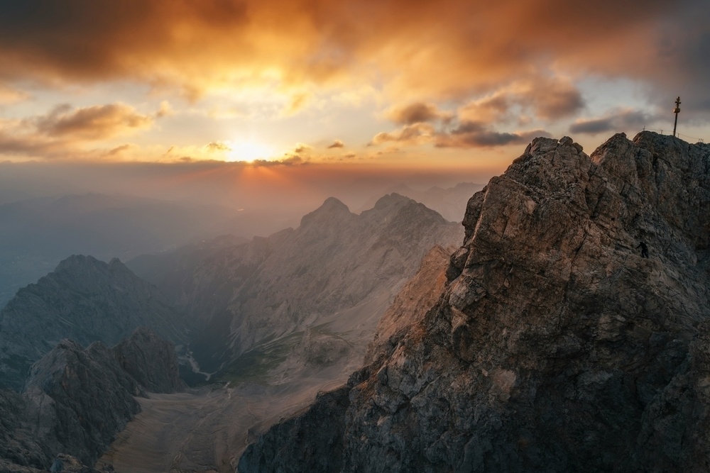 A dramatic sunrise casts a warm glow over rugged mountain peaks, highlighting a cross perched atop one of the rocky summits.