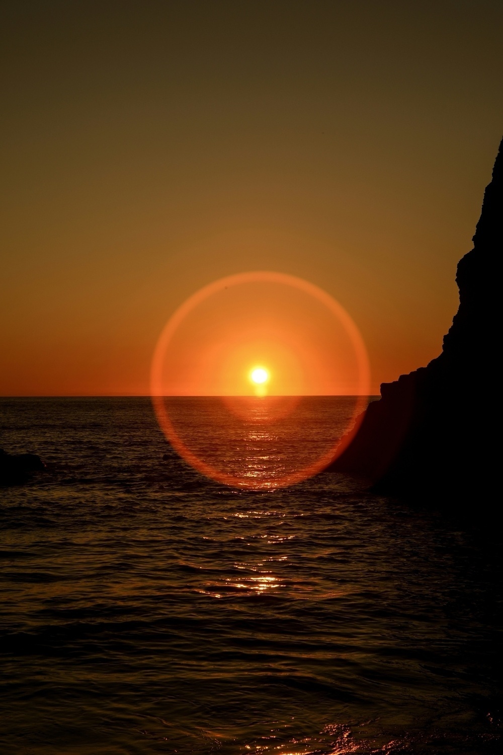 A sunset casts a warm glow over the ocean, with a prominent lens flare near the horizon.