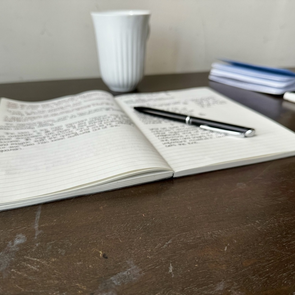 A notebook with handwritten text, a pen, and a cup sits on a wooden table.