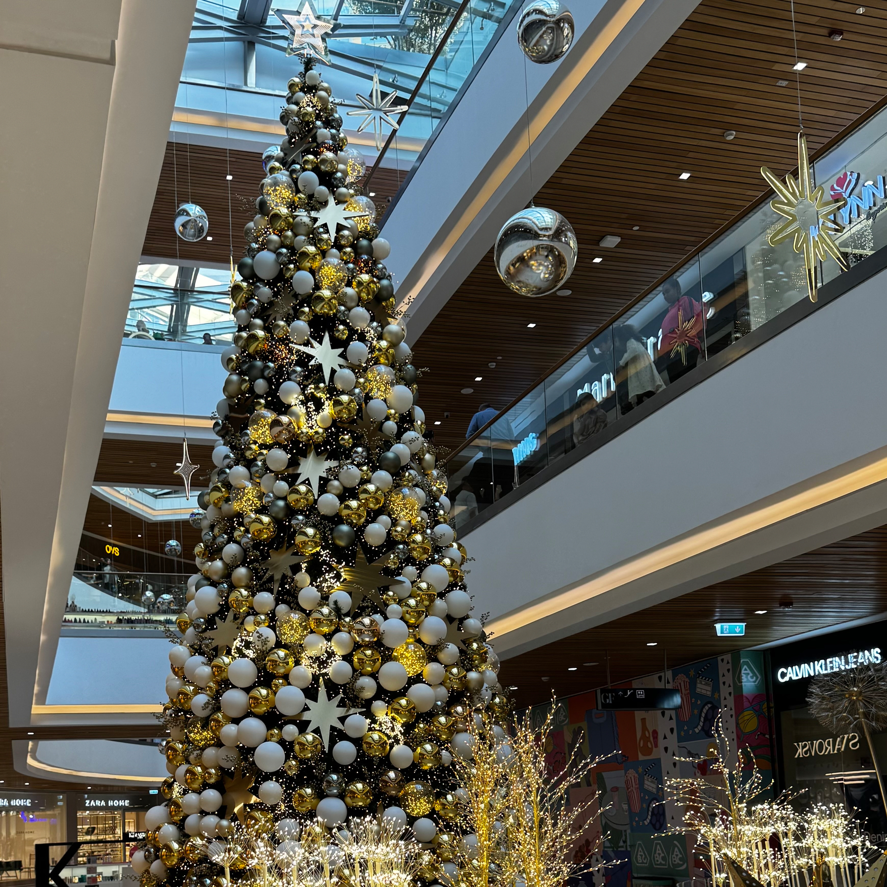 Auto-generated description: A large, beautifully decorated Christmas tree stands in a modern shopping mall with white and gold ornaments, surrounded by festive decorations.