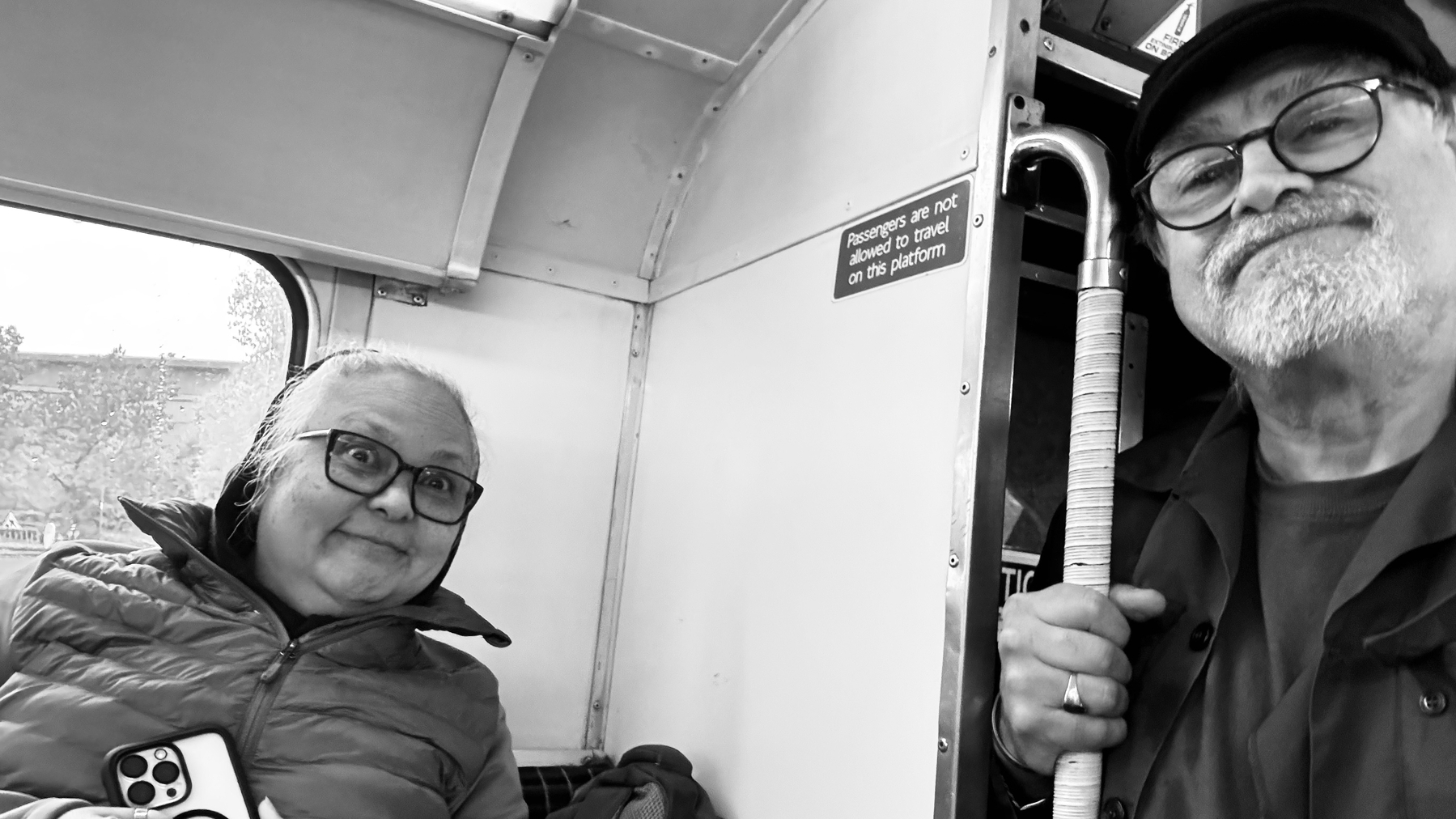 Two people are sitting on public transportation, one holding a phone and the other holding a walking cane, with both smiling at the camera.